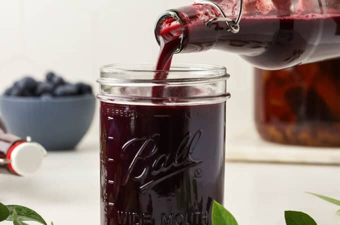 A jar cup with blueberry kombucha pouring into it, on a white countertop surrounded by fresh blueberry sprigs and a bowl of fresh blueberries in the background.