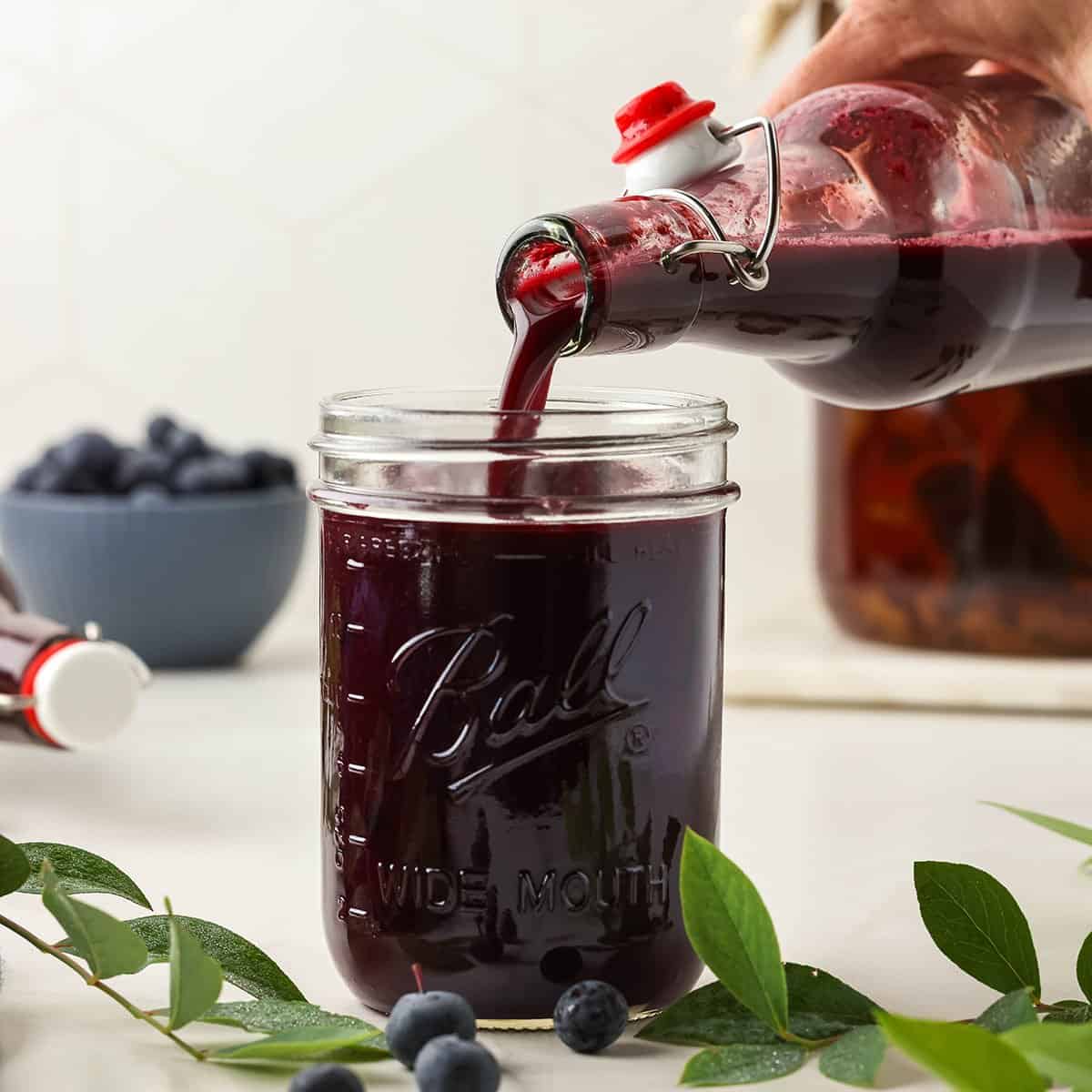 A jar cup with blueberry kombucha pouring into it, on a white countertop surrounded by fresh blueberry sprigs and a bowl of fresh blueberries in the background. 