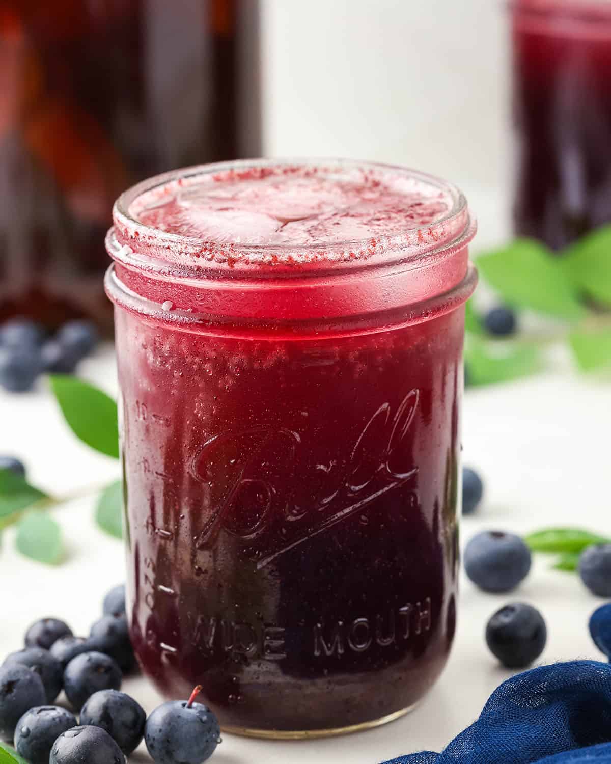 A jar of blueberry kombucha with ice in it, on a white countertop surrounded by fresh blueberries. 