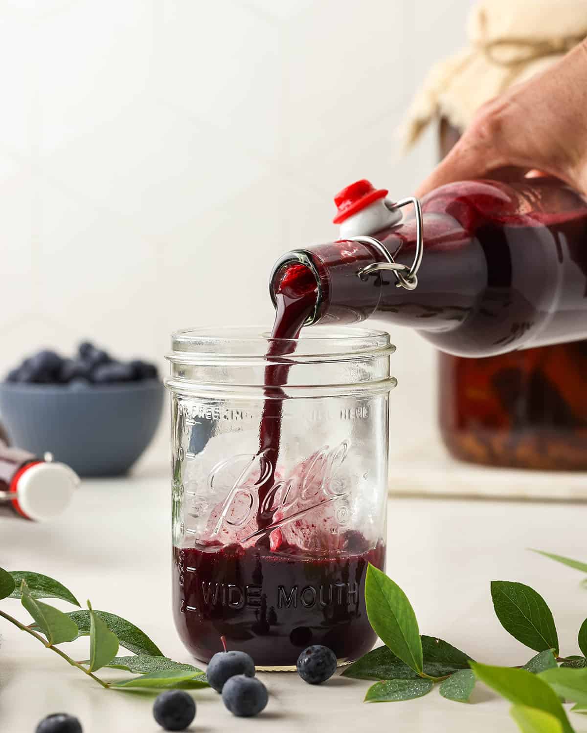 A bottle of blueberry kombucha pouring into a glass jar, on a white countertop surrounded by blueberry plants and berries. 