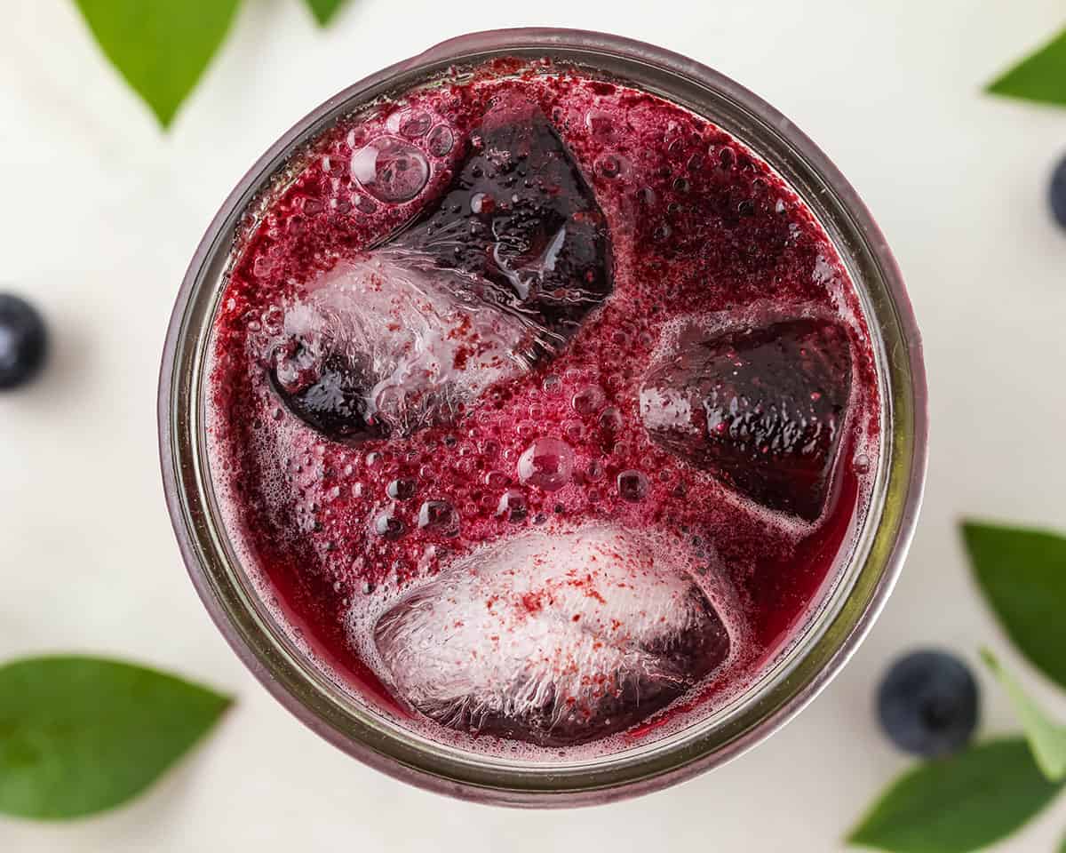 A jar of blueberry kombucha with ice on a white countertop surrounded by blueberry leaves and berries, top view. 