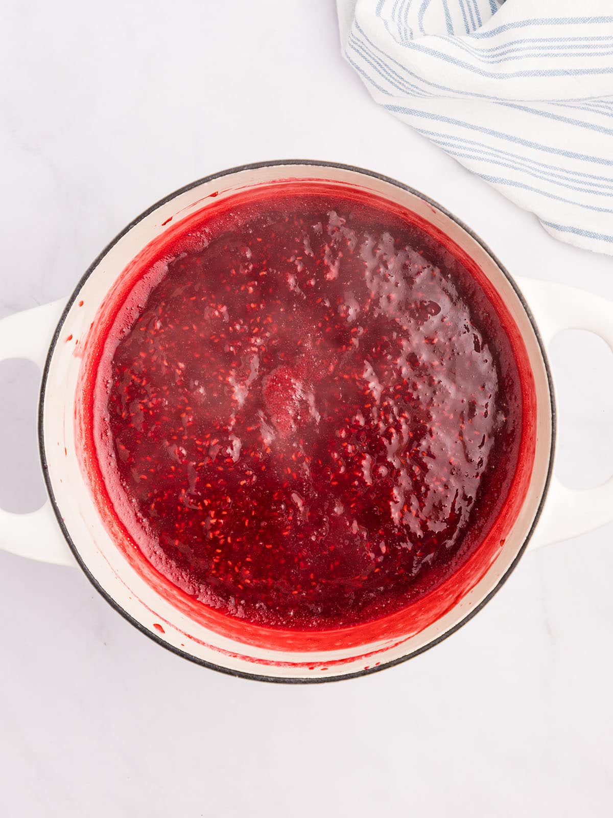 Cooked raspberry jam warm in a pot on a white surface, top view. 