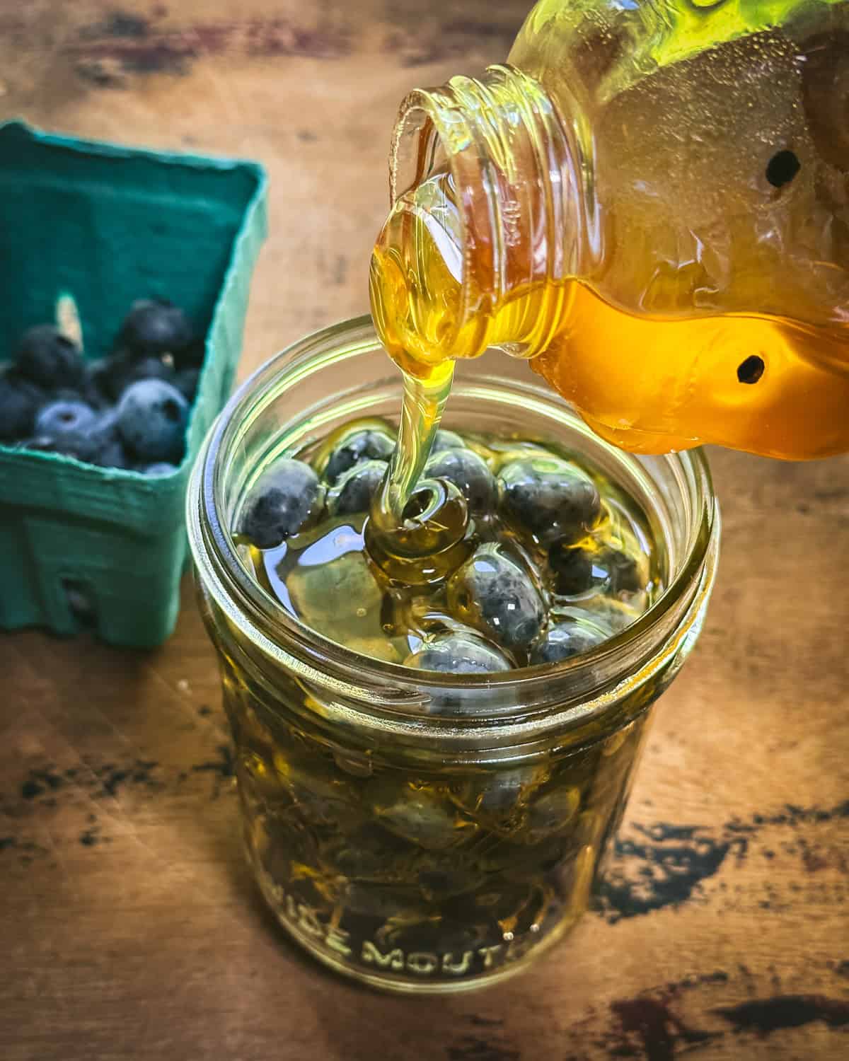 A jar of blueberries with honey pouring in, on a wood surface. 