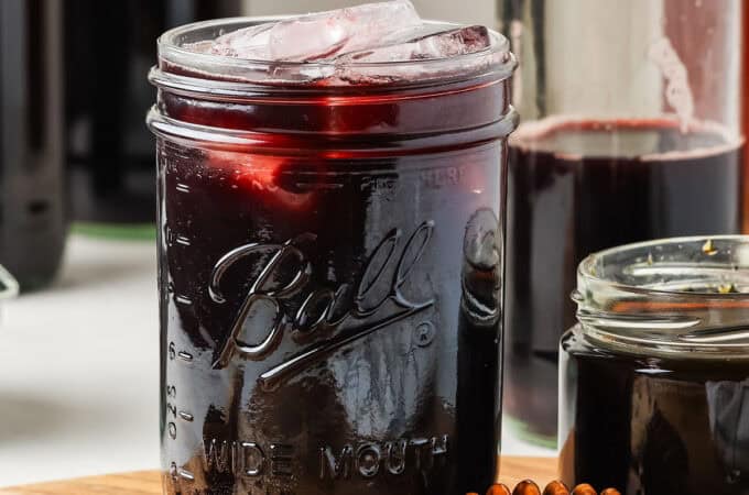 A jar of dark elderberry kombucha with ice, on a wood cutting board, surrounded by fresh elderberries, a honey wand, and elderberry syrup.