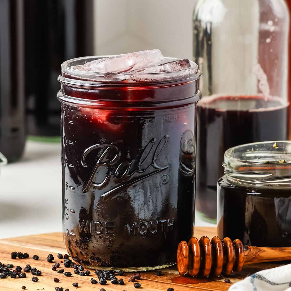 A jar of dark elderberry kombucha with ice, on a wood cutting board, surrounded by fresh elderberries, a honey wand, and elderberry syrup.