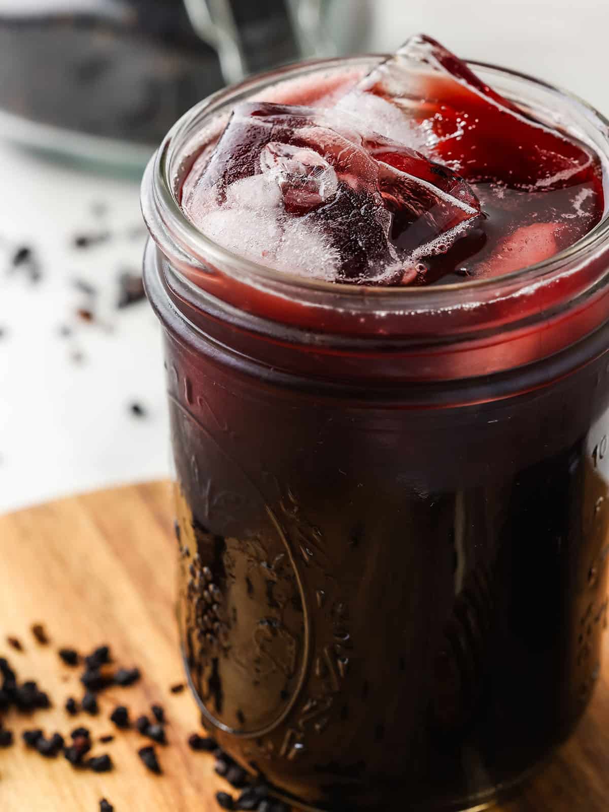 Elderberry kombucha in a glass jar with ice on a wood cutting board. 