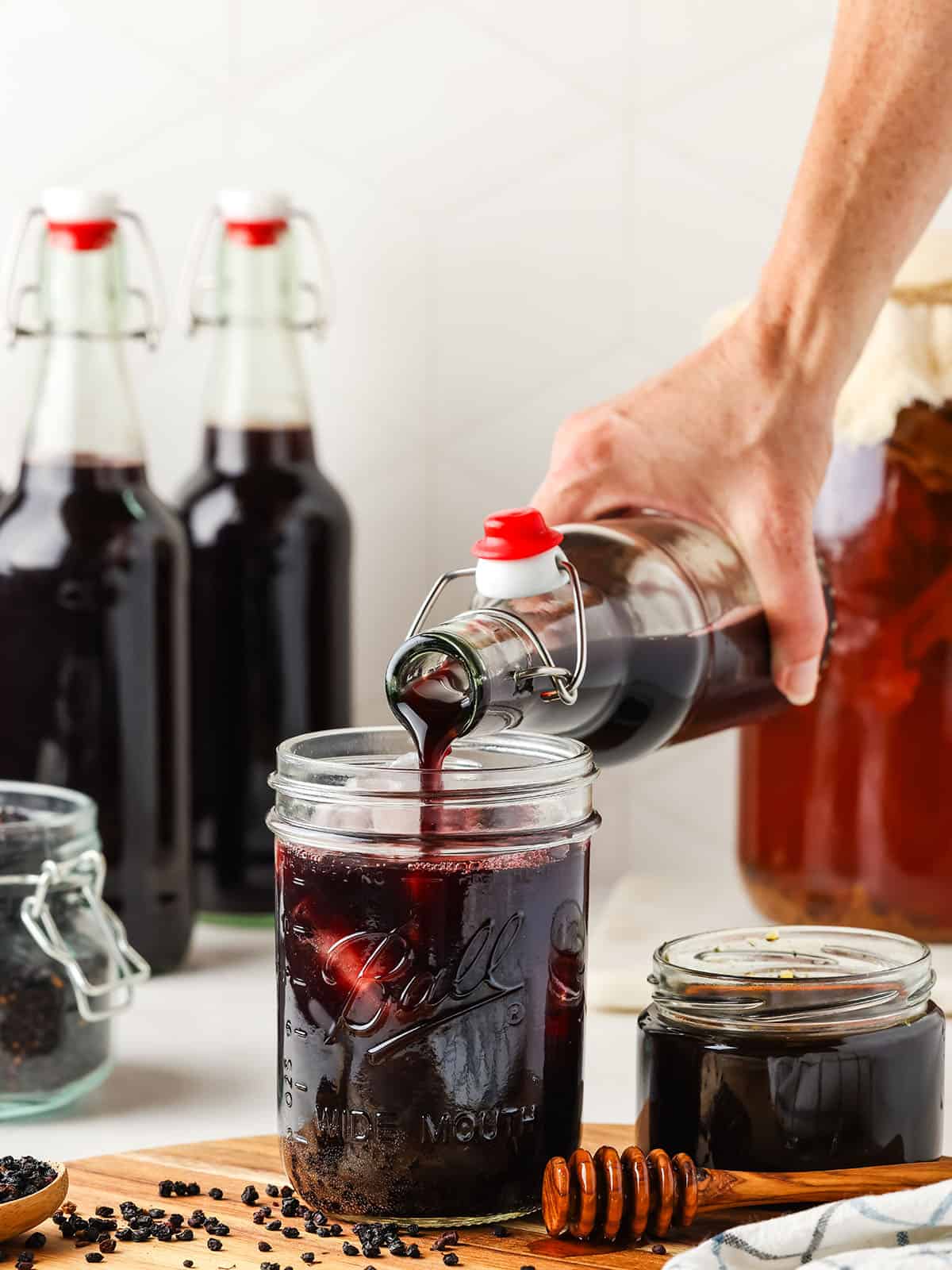 Elderberry kombucha pouring into a glass jar over ice with more bottles in the background. 