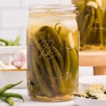 A jar of fermented dilly beans on a white counter surrounded by fresh green beans.