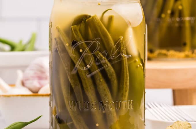 A jar of fermented dilly beans on a white counter surrounded by fresh green beans.