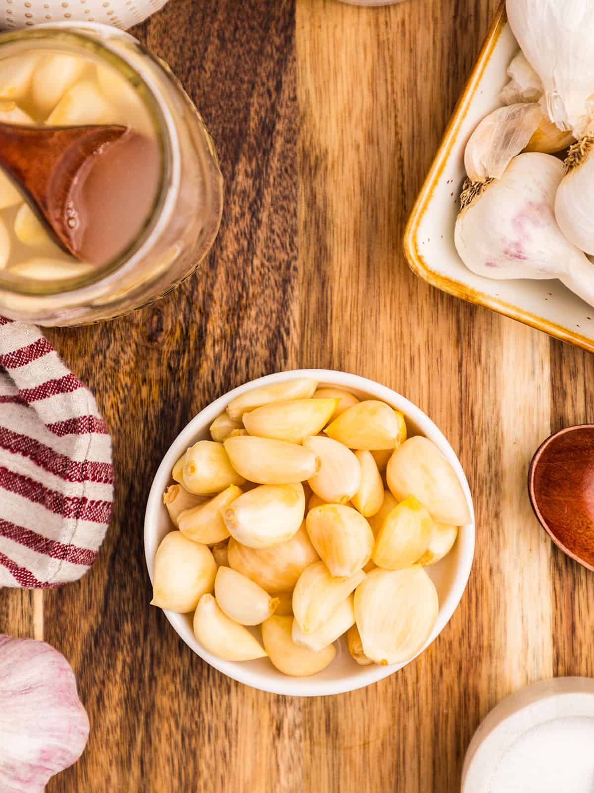 A bowl of fermented garlic on a wood surface, surrounded by a jar of fermented garlic. 