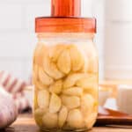 A jar of fermenting garlic with an orange airlock lid on top, on a wood surface surrounded by fresh garlic.