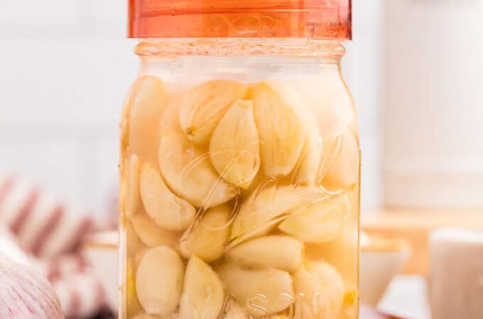 A jar of fermenting garlic with an orange airlock lid on top, on a wood surface surrounded by fresh garlic.