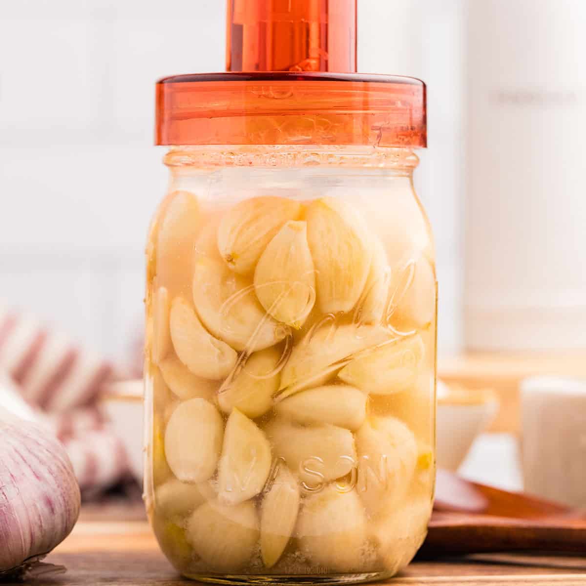 A jar of fermenting garlic with an orange airlock lid on top, on a wood surface surrounded by fresh garlic. 