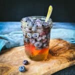 A jar of honey with blueberries in it floating to the top. On a wood cutting board with blue fabric in the background and loose fresh blueberries around.