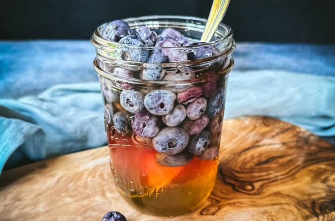 A jar of honey with blueberries in it floating to the top. On a wood cutting board with blue fabric in the background and loose fresh blueberries around.