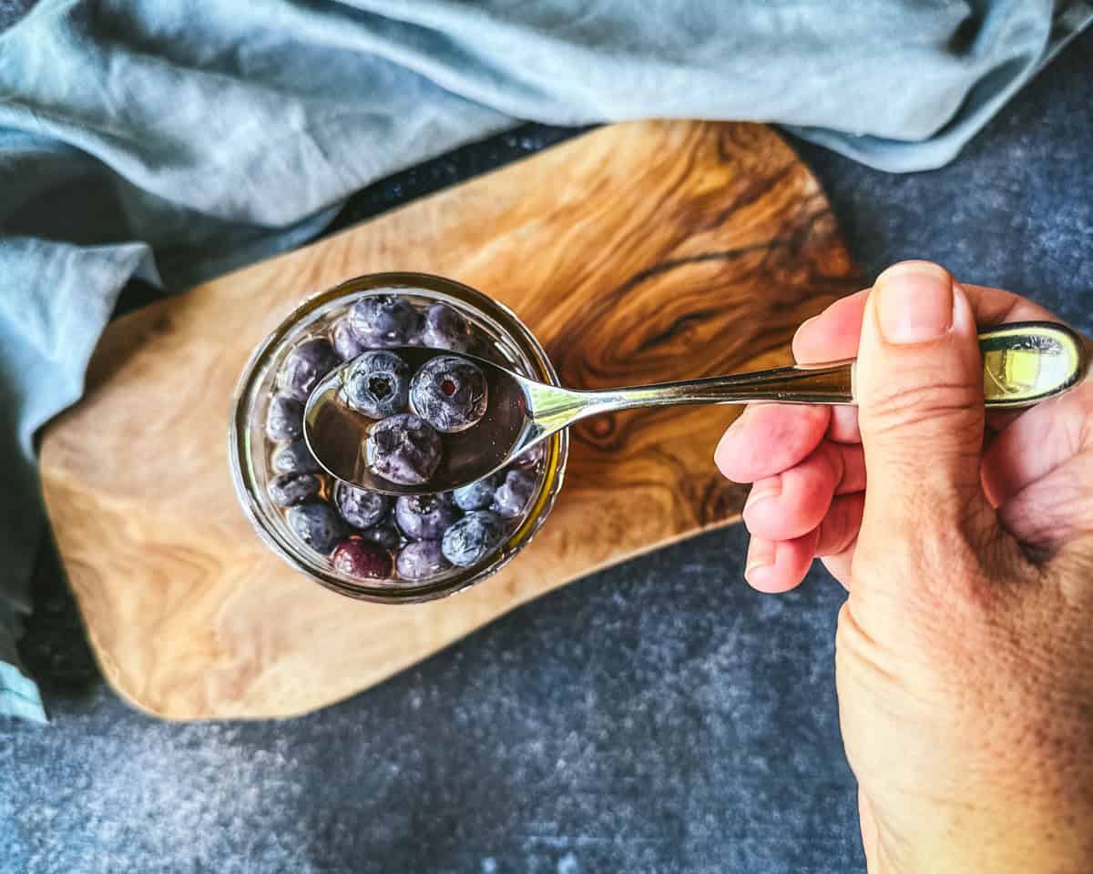 Fermented honey blueberries on a spoon lifting up out of the jar, on a wood surface. Top view. 