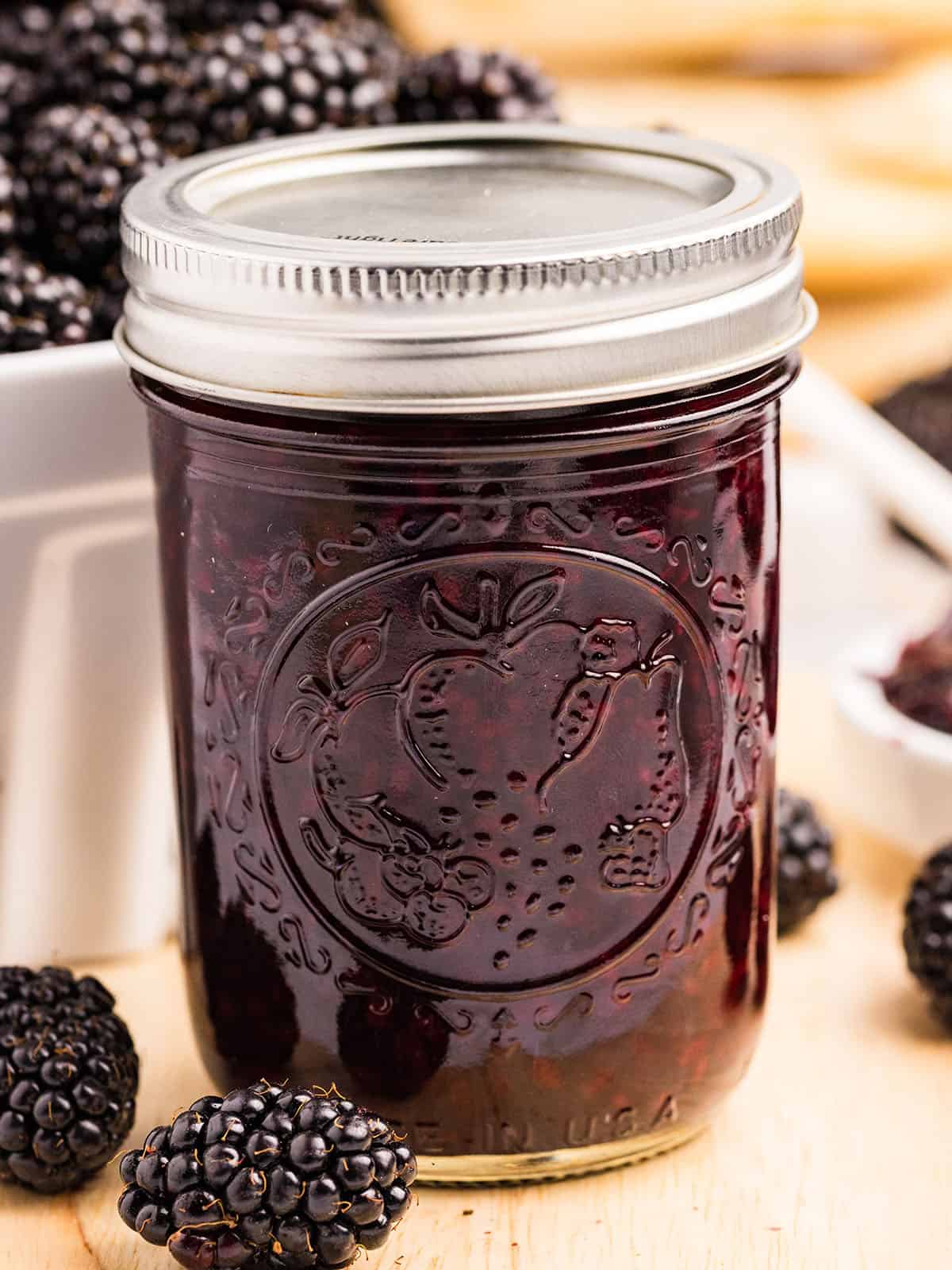 A jar of homemade blackberry jam with fresh blackberries surrounding. 