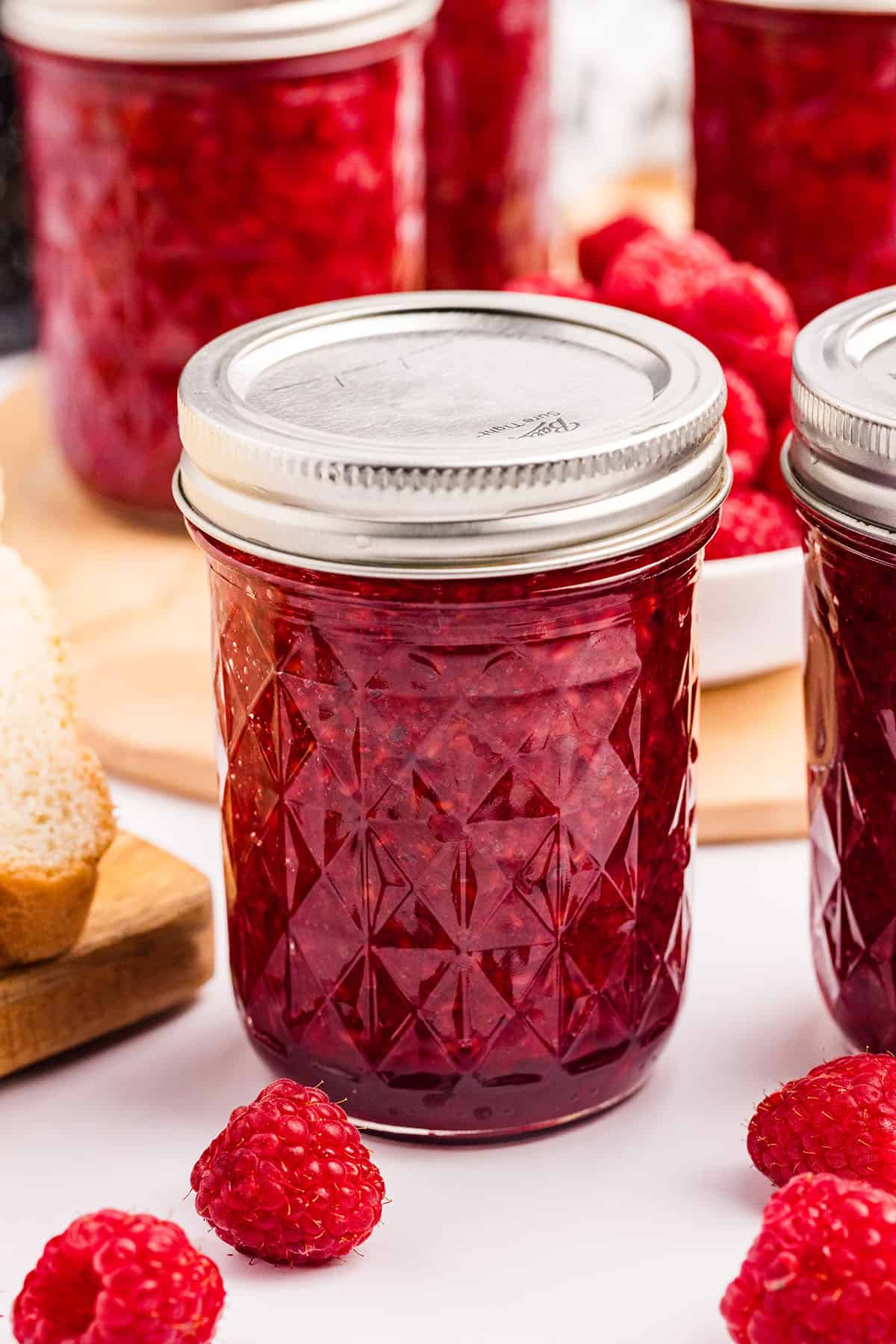 A canned jar of raspberry jam with other jars and fresh raspberries surrounding. 