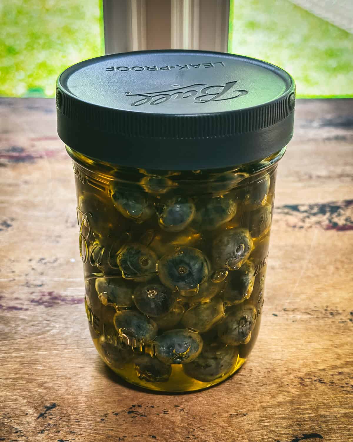 A jar of blueberries in honey with a plastic lid, on a wood surface. 