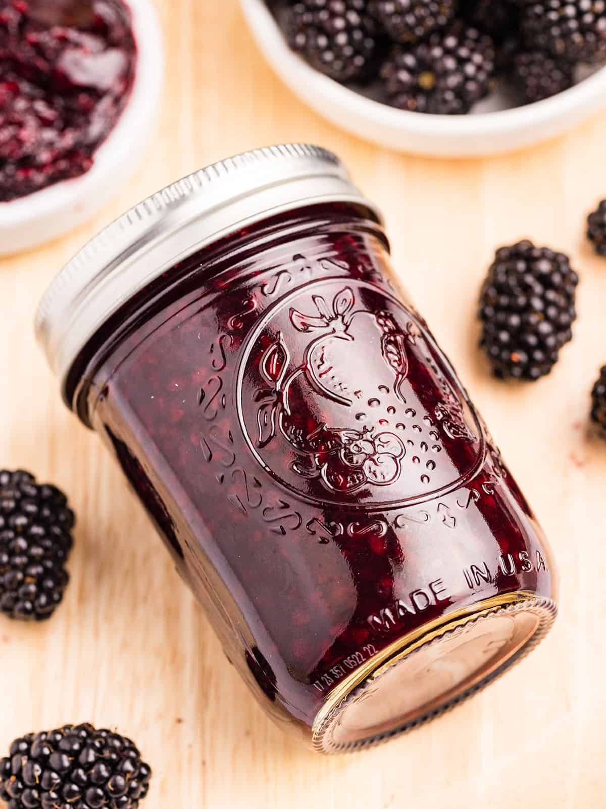 A jar of blackberry jam on on its side on a light wood surface surrounded by fresh blackberries. 