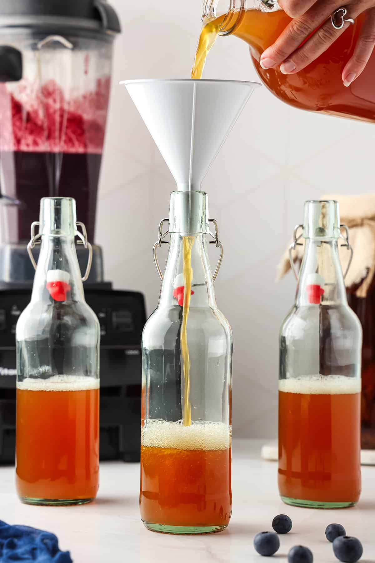 3 flip top bottles halfway filled with kombucha starter, one with a funnel on top with kombucha being poured in. 