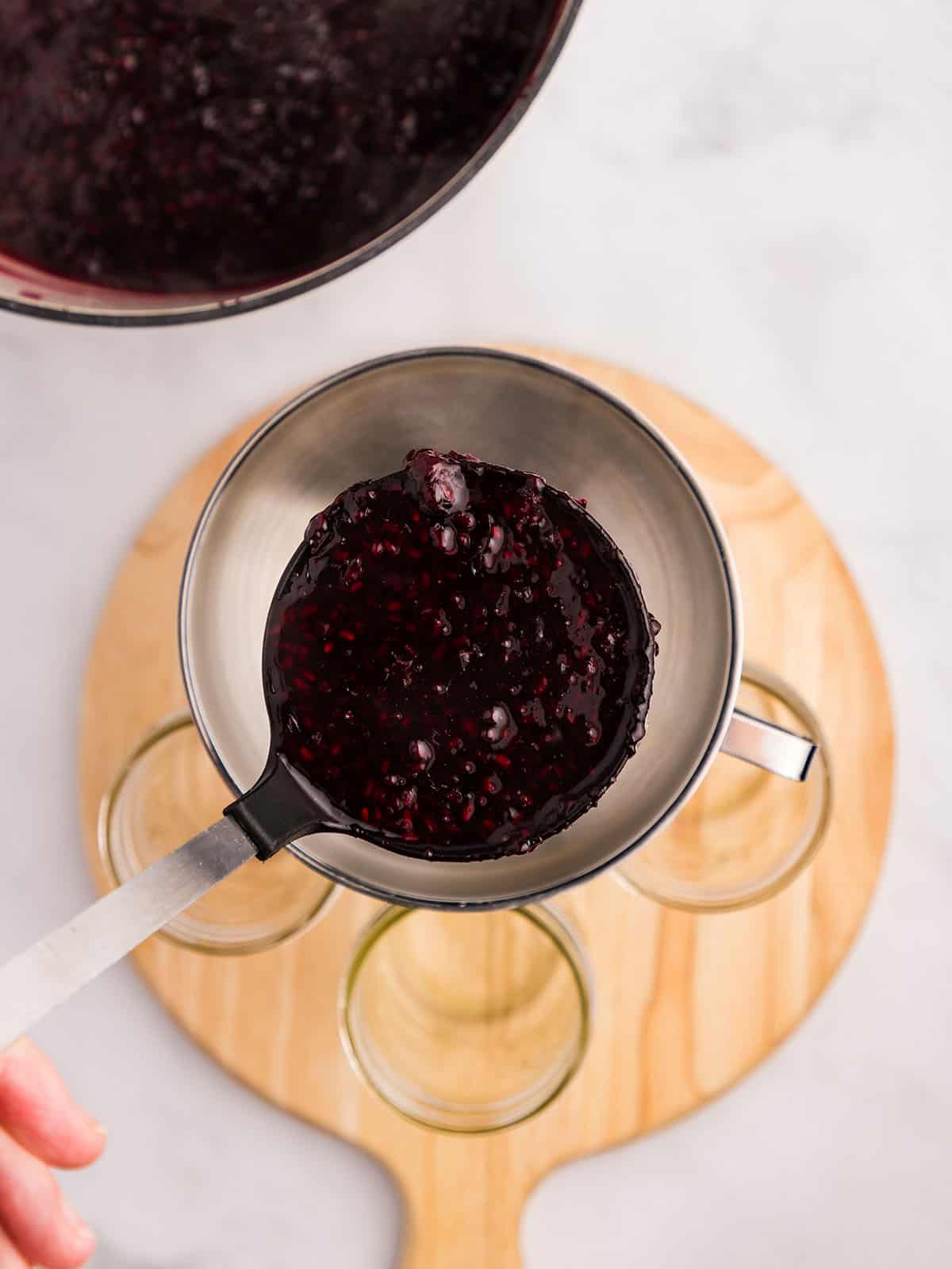 A ladle pouring blackberry jam into jars on a circular wood cutting board, top view. 