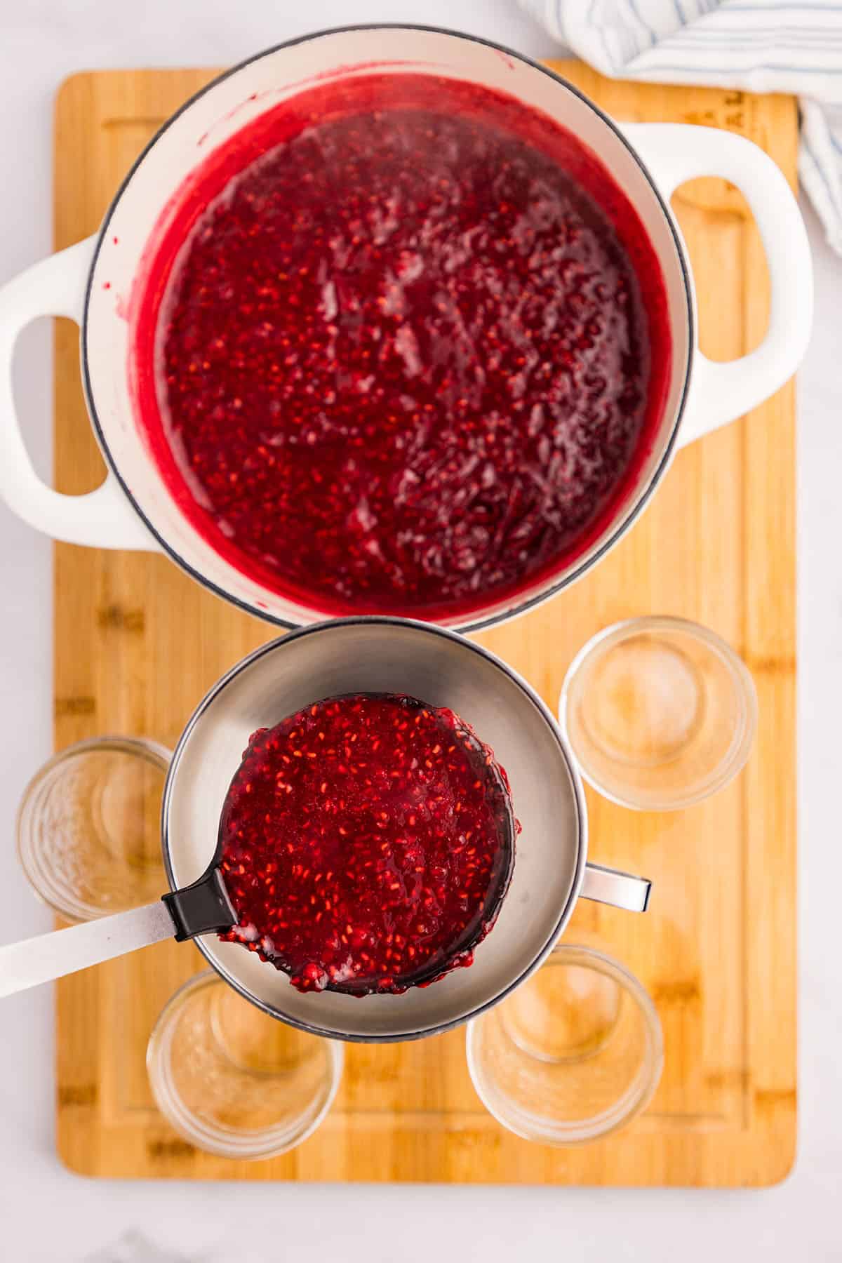 A ladle pouring raspberry jam into a jar with a funnel, top view. 