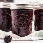 Jars of dark purple blackberry jam on a cloth with fresh blackberries surrounding.