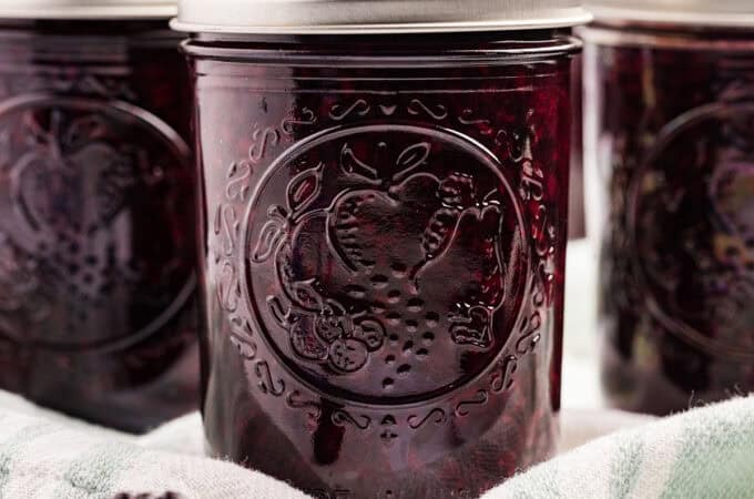 Jars of dark purple blackberry jam on a cloth with fresh blackberries surrounding.