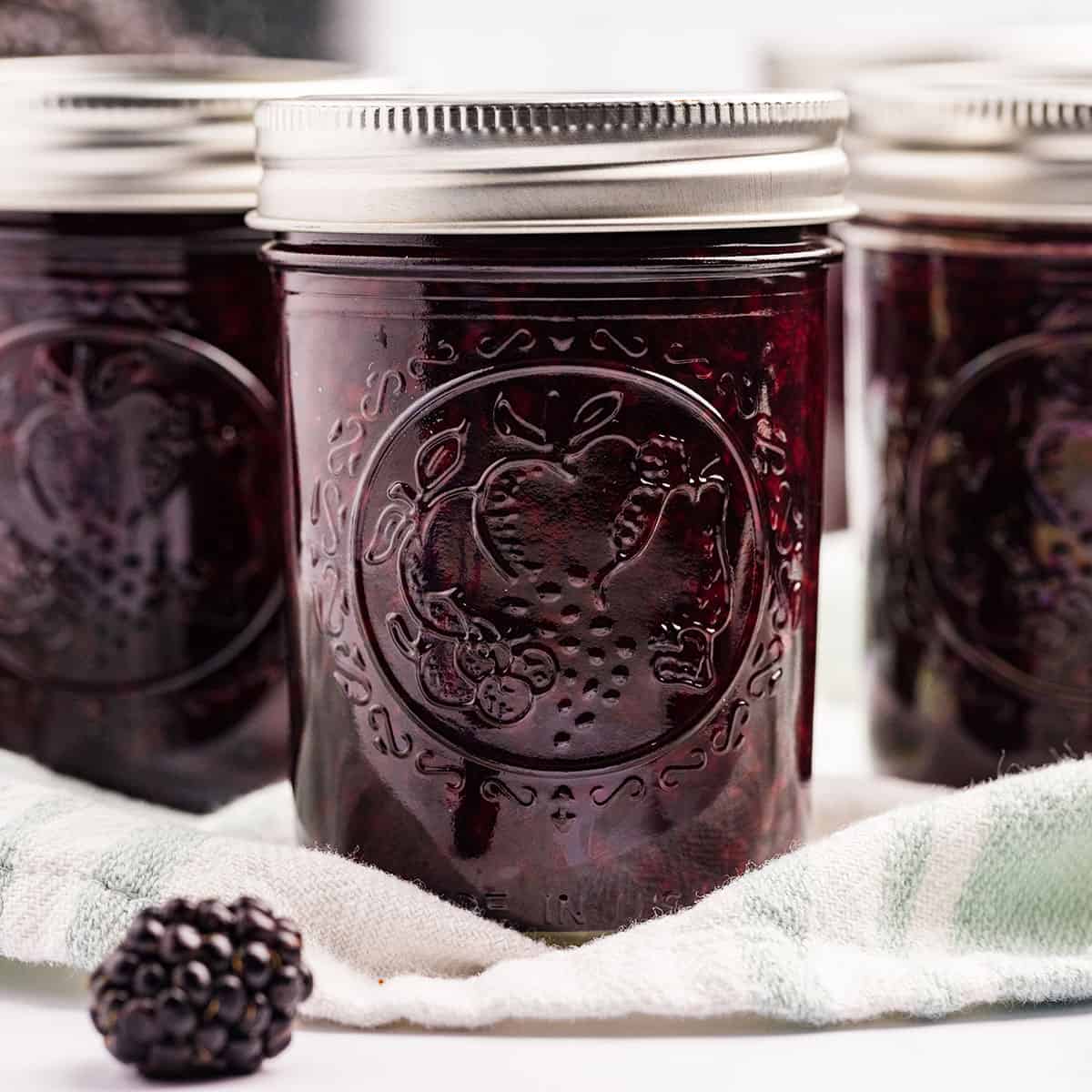 Jars of dark purple blackberry jam on a cloth with fresh blackberries surrounding. 