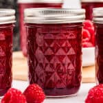 A jar of deep red raspberry jam, on a white countertop with fresh raspberries surrounding.