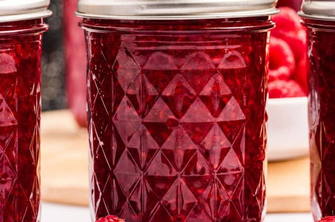 A jar of deep red raspberry jam, on a white countertop with fresh raspberries surrounding.