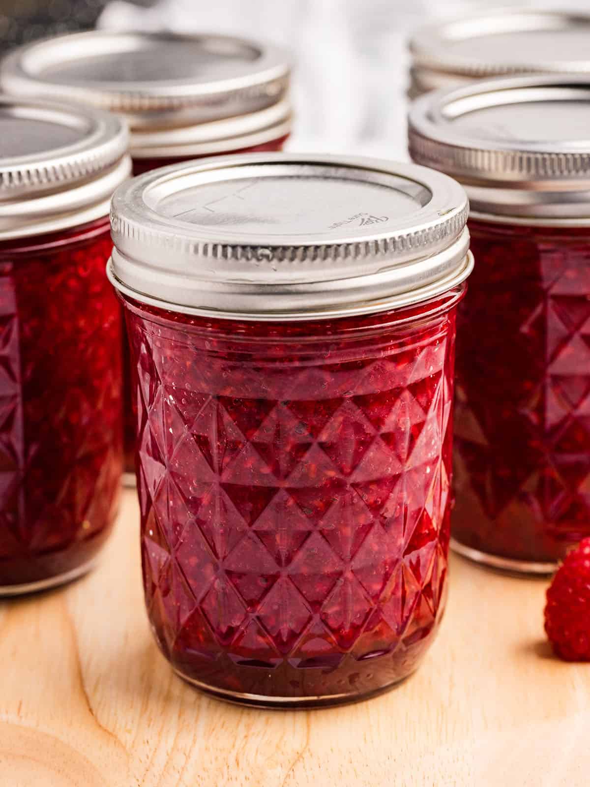 A jar of finished and canned raspberry jam on a wood surface, with other jars of raspberry jams in the background. 