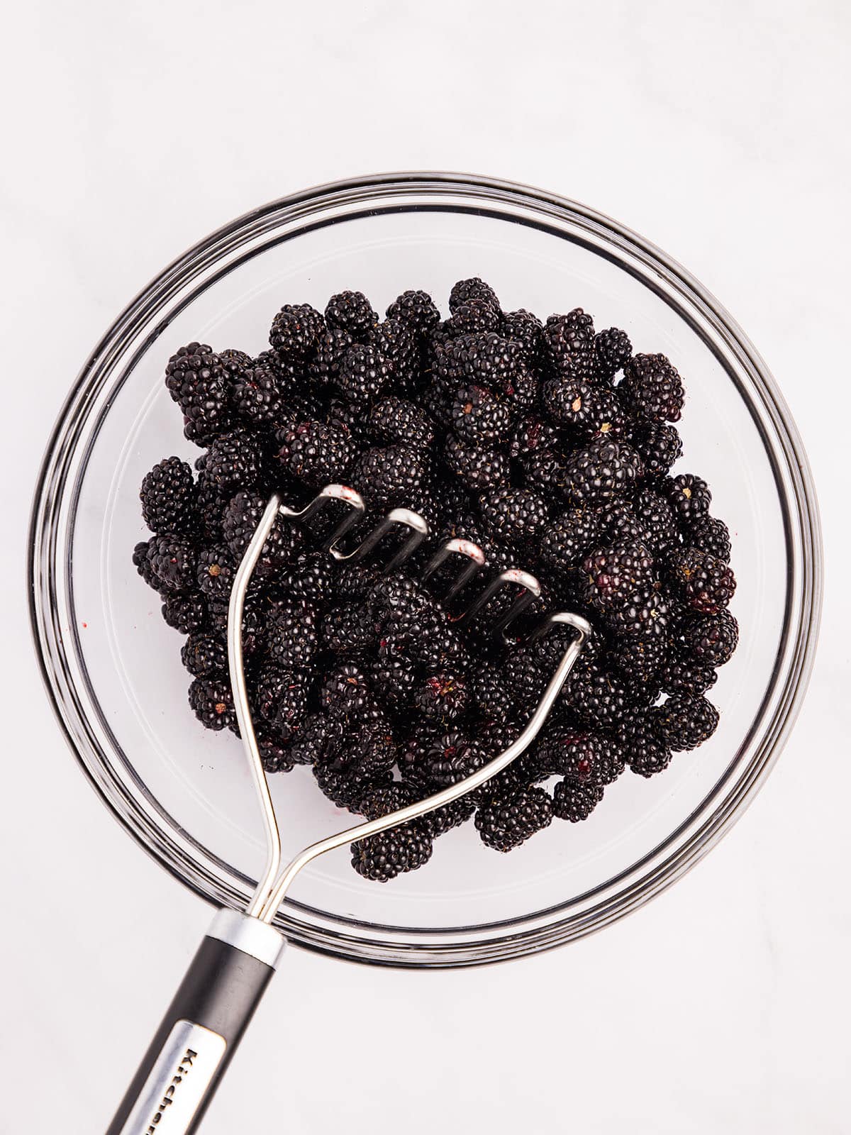 Blackberries being mashed with a potato masher in a bowl, top view. 