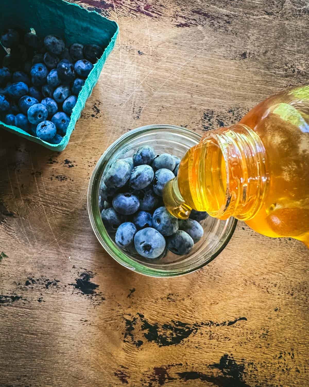 A jar with blueberries at the bottom, and honey pouring in, on a wood surface, with a pint of blueberries to the side. Top view. 