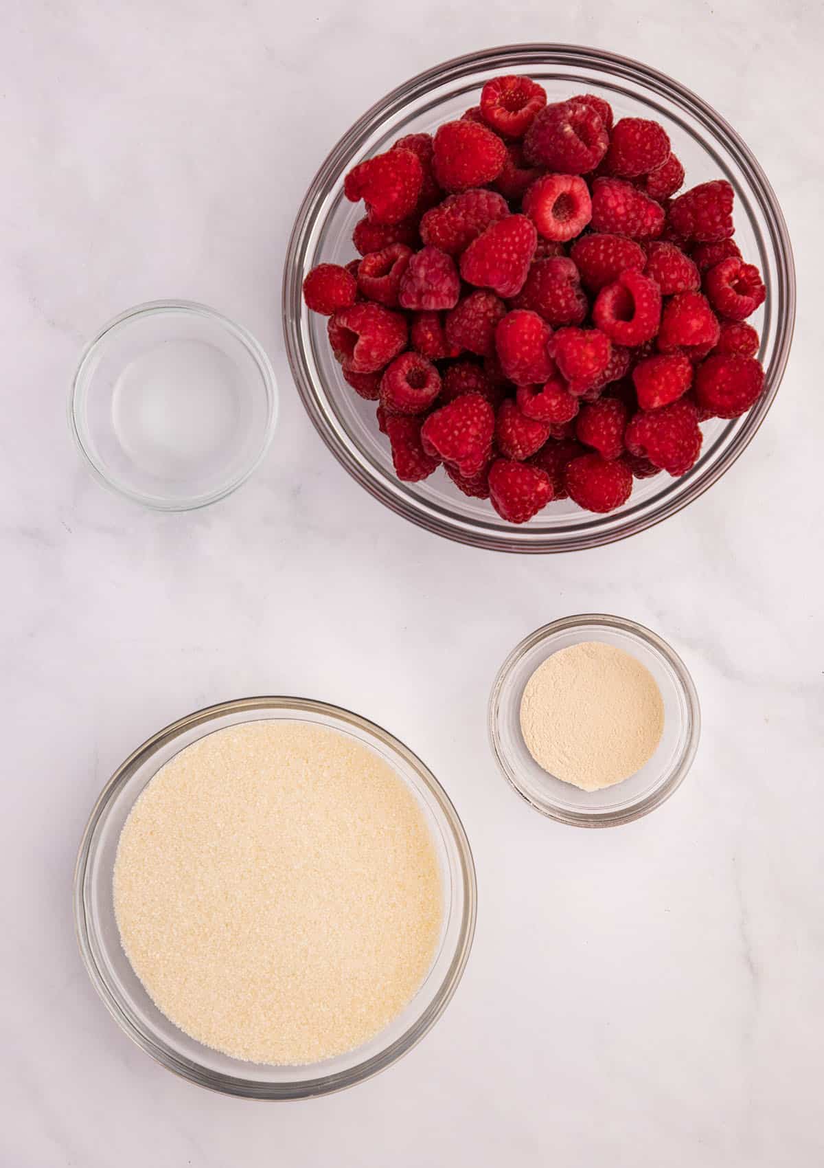 Bowls of raspberry jam ingredients on a white counter, top view.