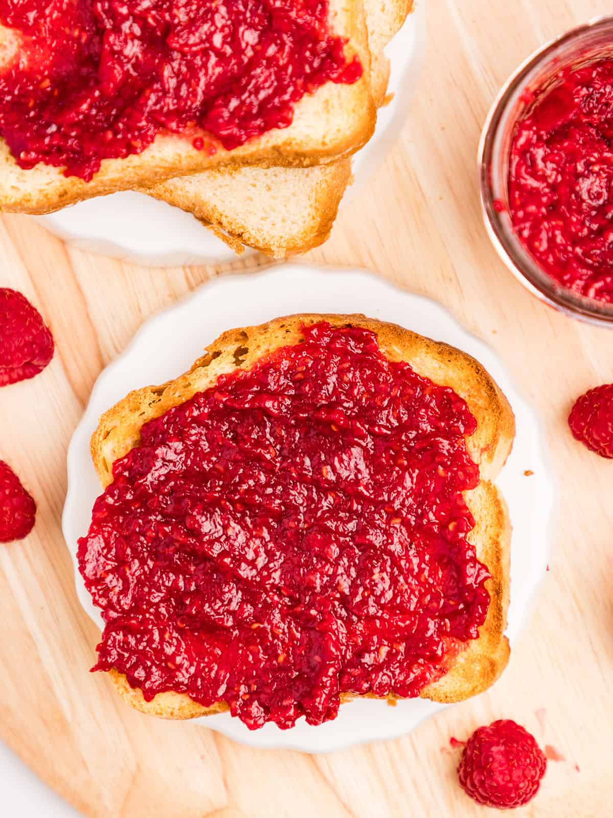 A plate with toast spread with low sugar raspberry jam on it, sitting on a light wood surface surrounded by fresh raspberries and a jar of raspberry jam. Top view. 