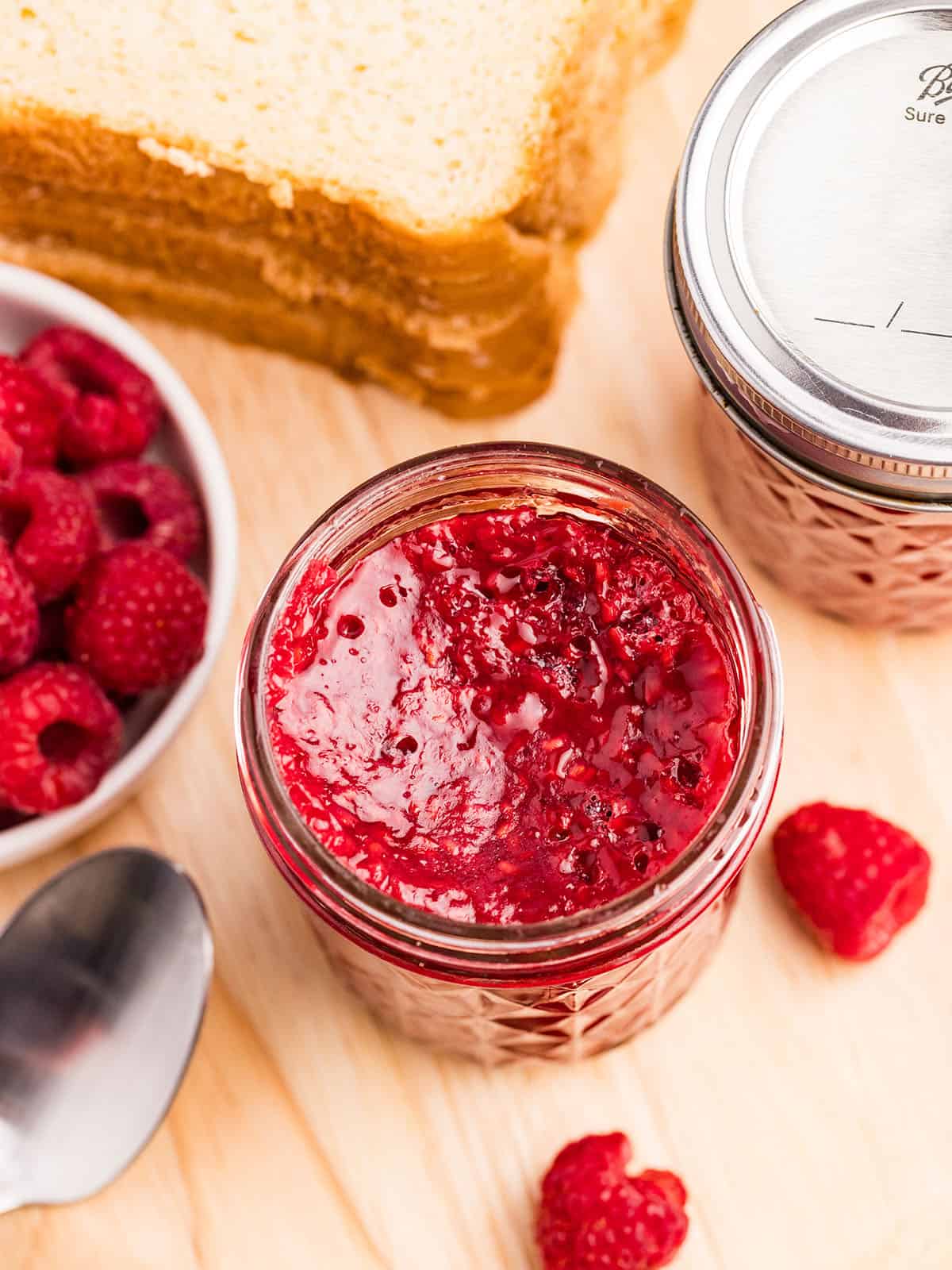 An open jar of raspberry jam on a wood cutting board surrounded by a bowl of fresh raspberries, and bread. 