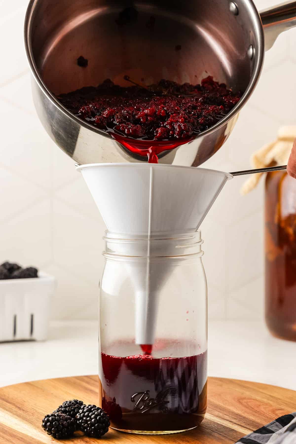 A pot pouring blackberry puree through a funnel into a jar, on a wood surface. 