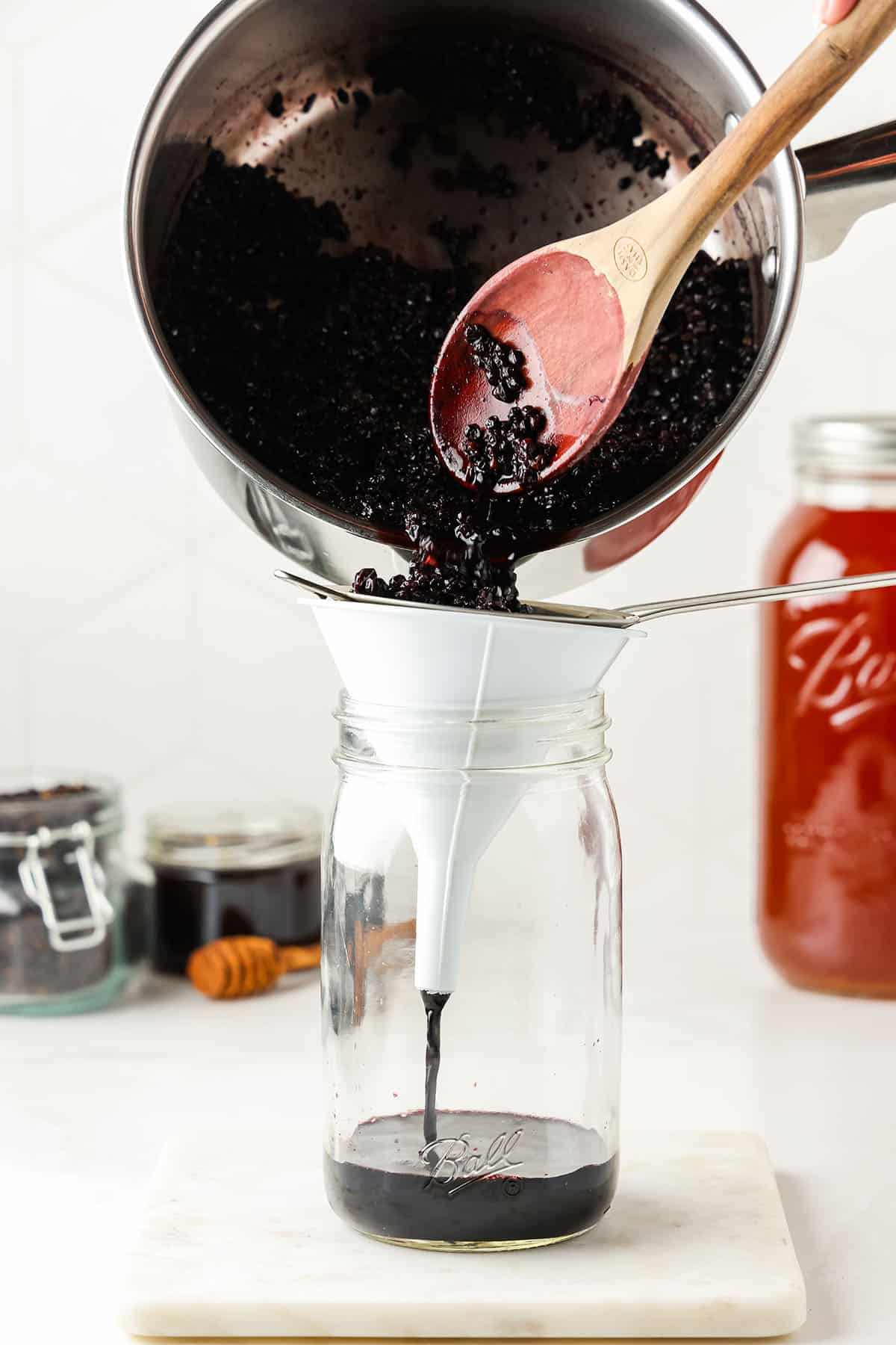 The pot of boiled elderberries pouring through a funnel and sieve and into a jar. 