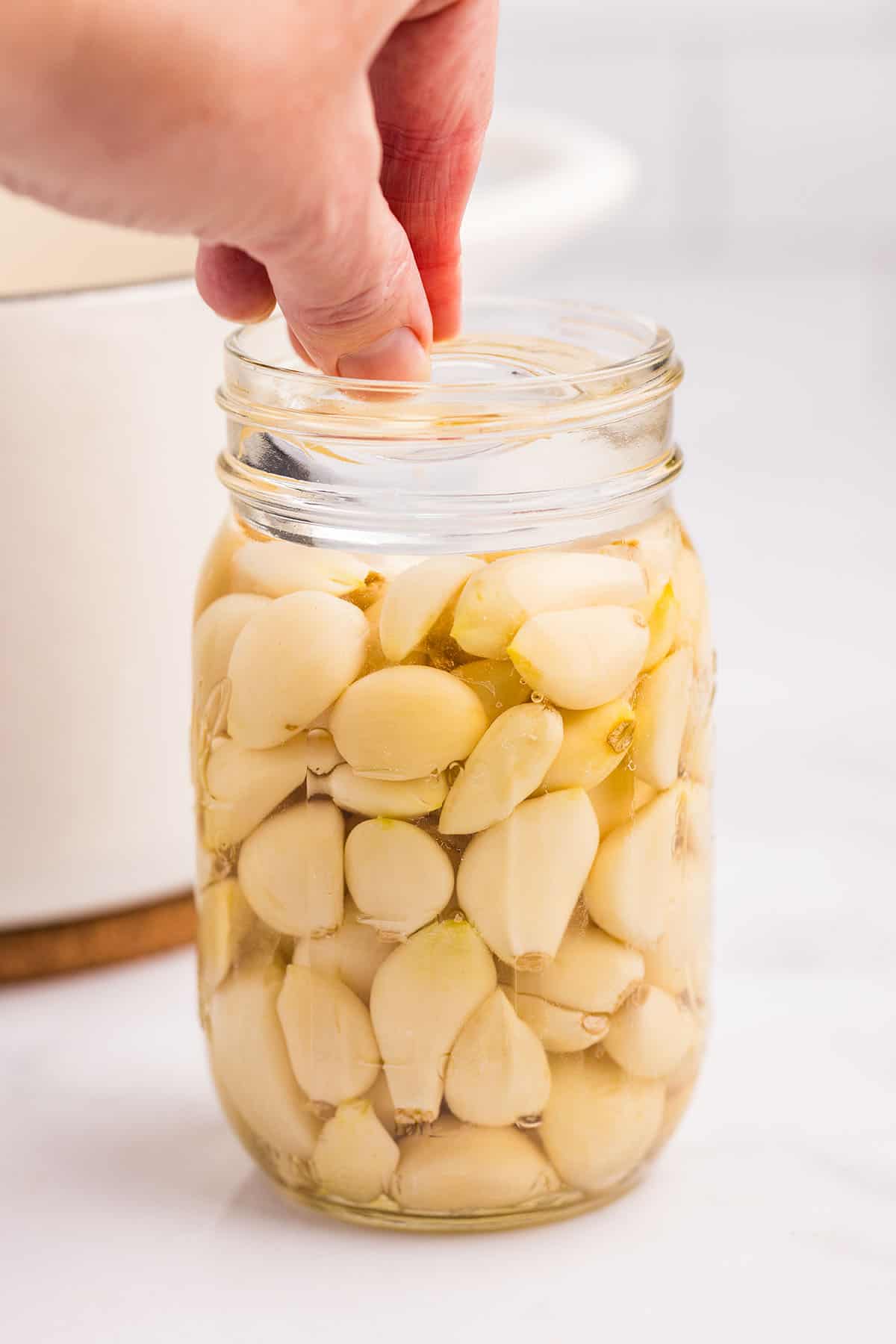 A jar of garlic and brine with a hand placing a fermentation weight on top. 