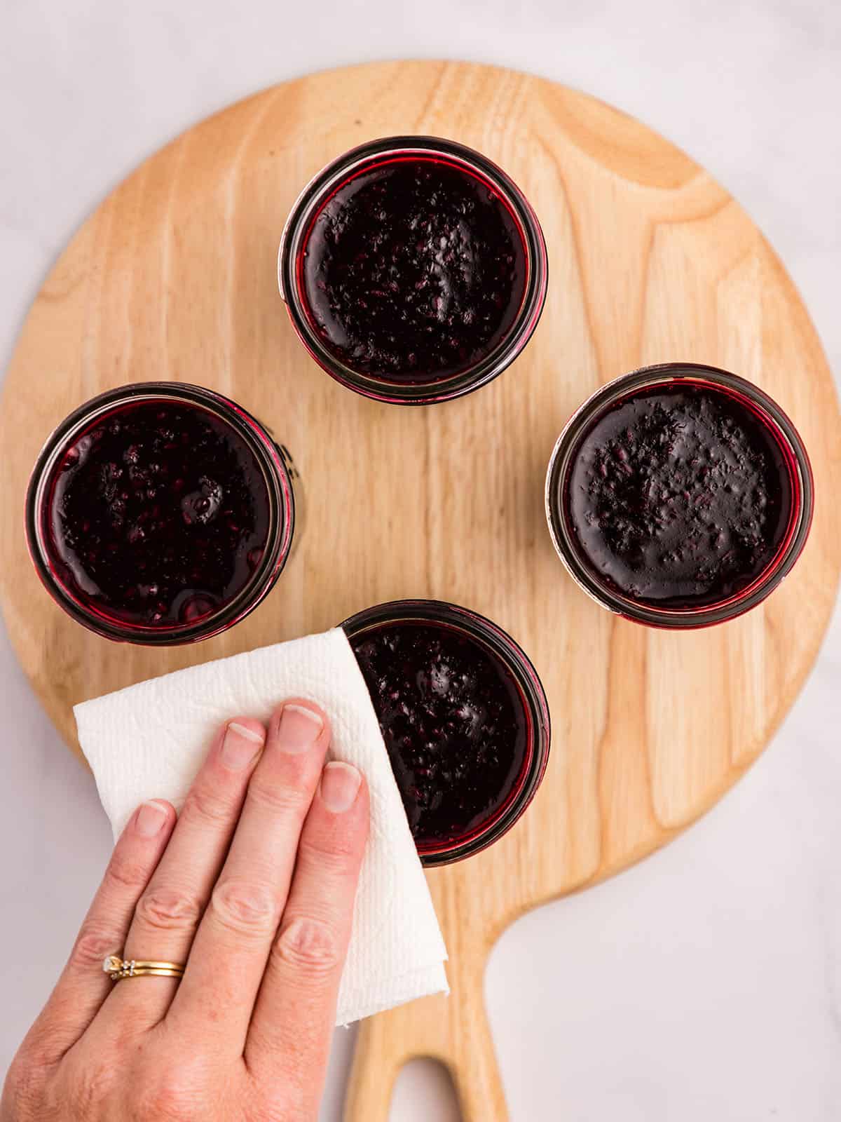 4 jars of blackberry jam on a circular wood cutting board, with a hand wiping the rim with a white cloth. Top view. 