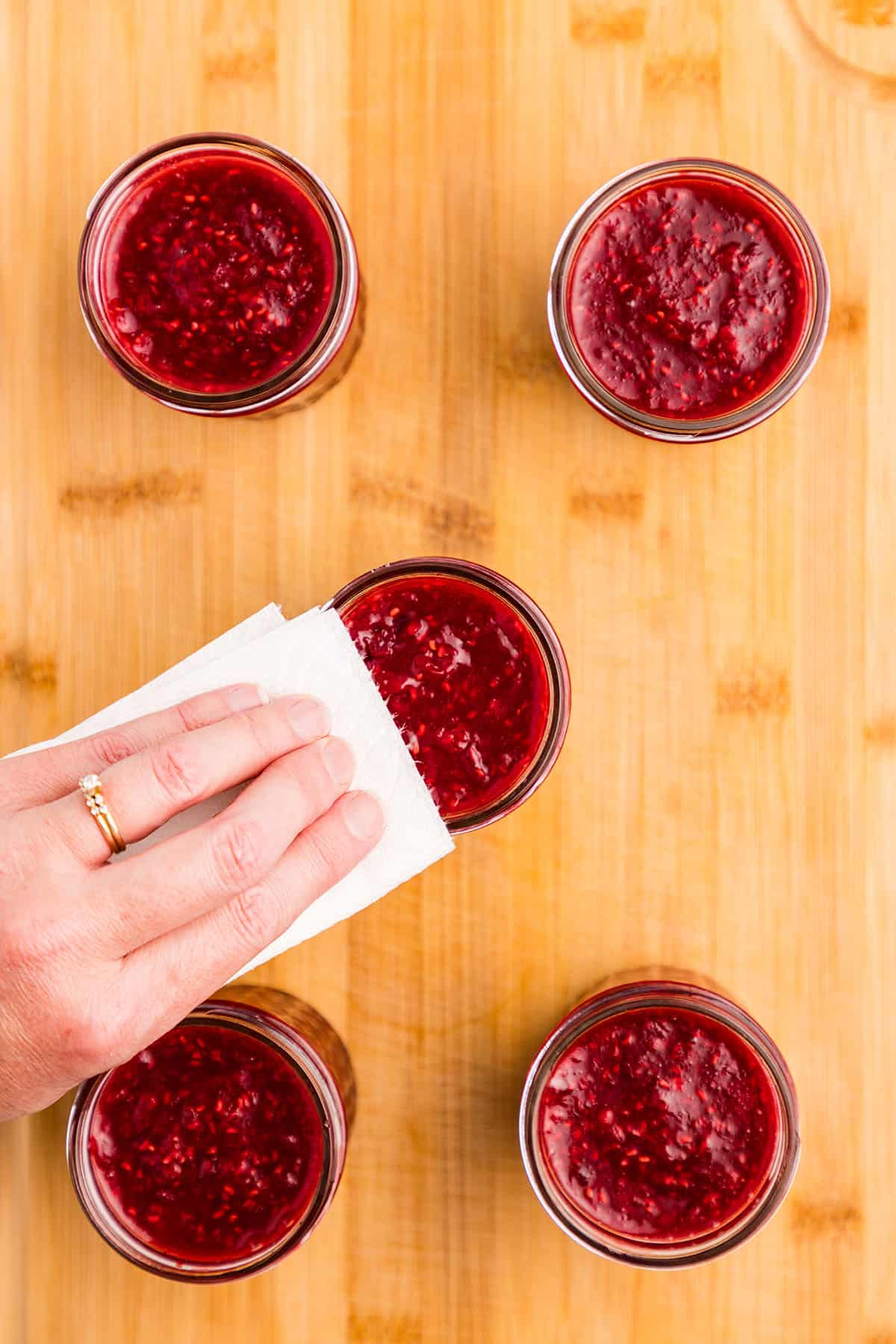 5 jars filled with raspberry jam on a wood surface, with a hand wiping the rim of the center jar. 