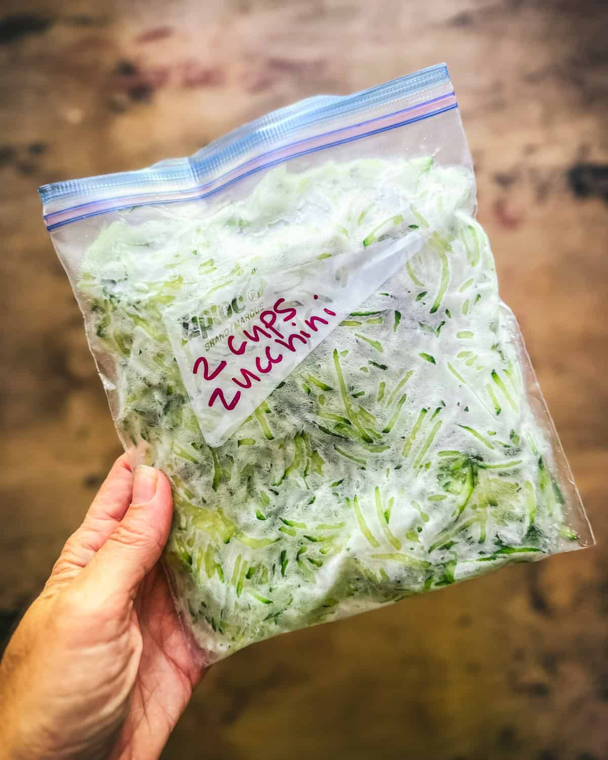 A hand holding a bag of frozen shredded zucchini over a wood surface. 