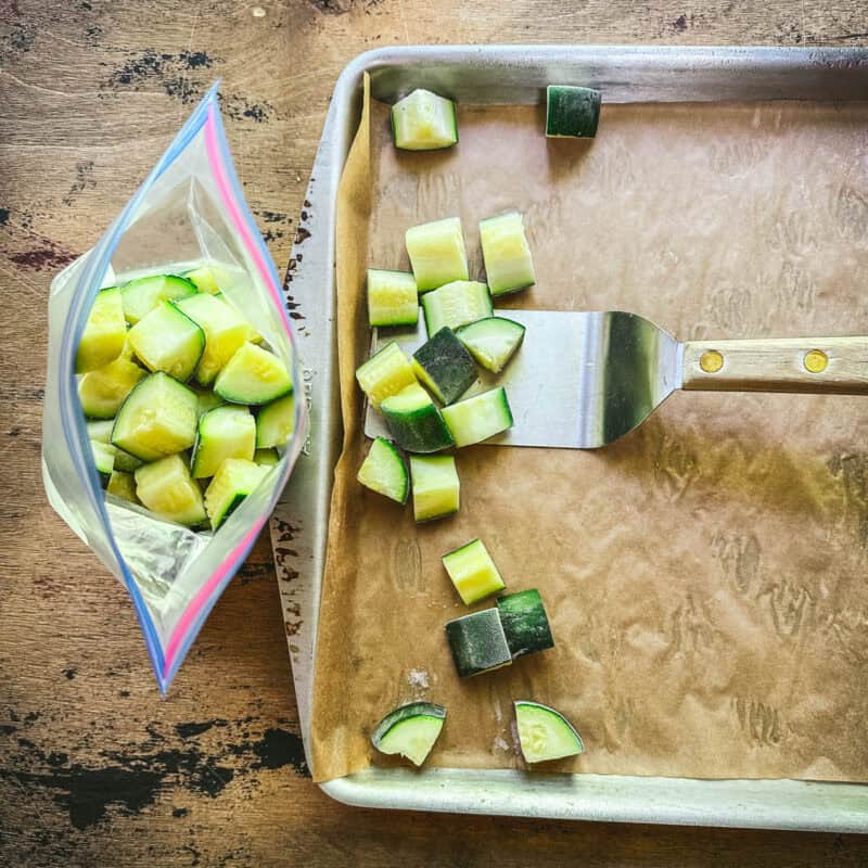 A spatula scooping frozen zucchini cubes from a sheet pan into a bag.