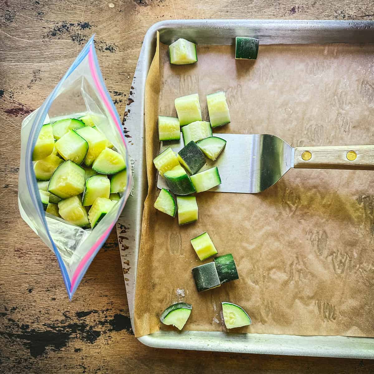 A spatula scooping frozen zucchini cubes from a sheet pan into a bag. 