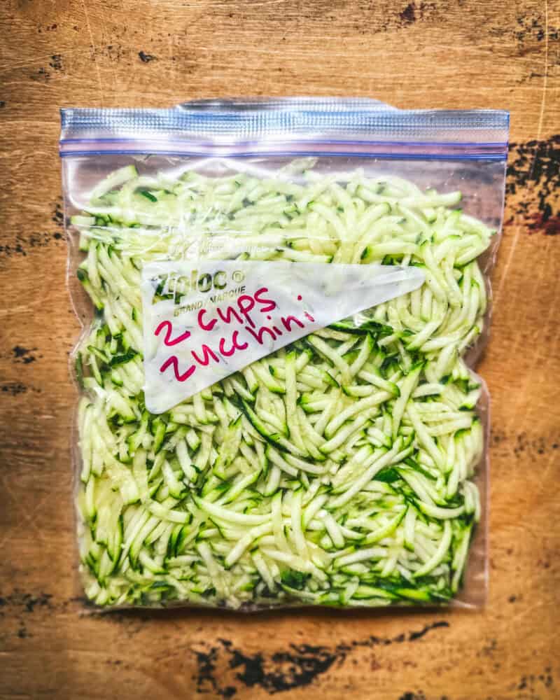 Shredded zucchini in a zip-top freezer bag on a wood surface.