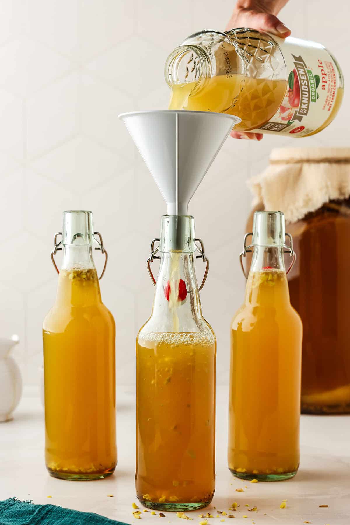 3 flip top bottles of kombucha, one with a funnel with apple juice pouring into it. 