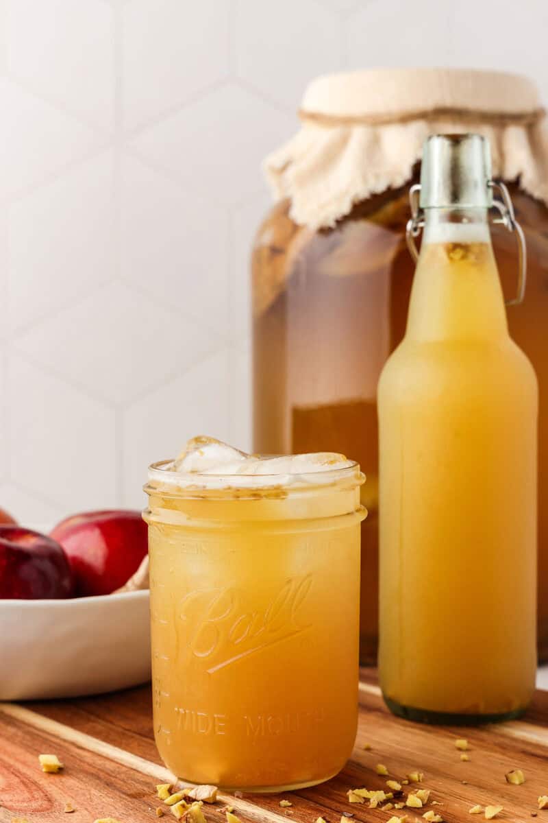 A glass with ice and kombucha next to a bottle of kombucha and a gallon of basic kombucha with cheesecloth secured to the top.