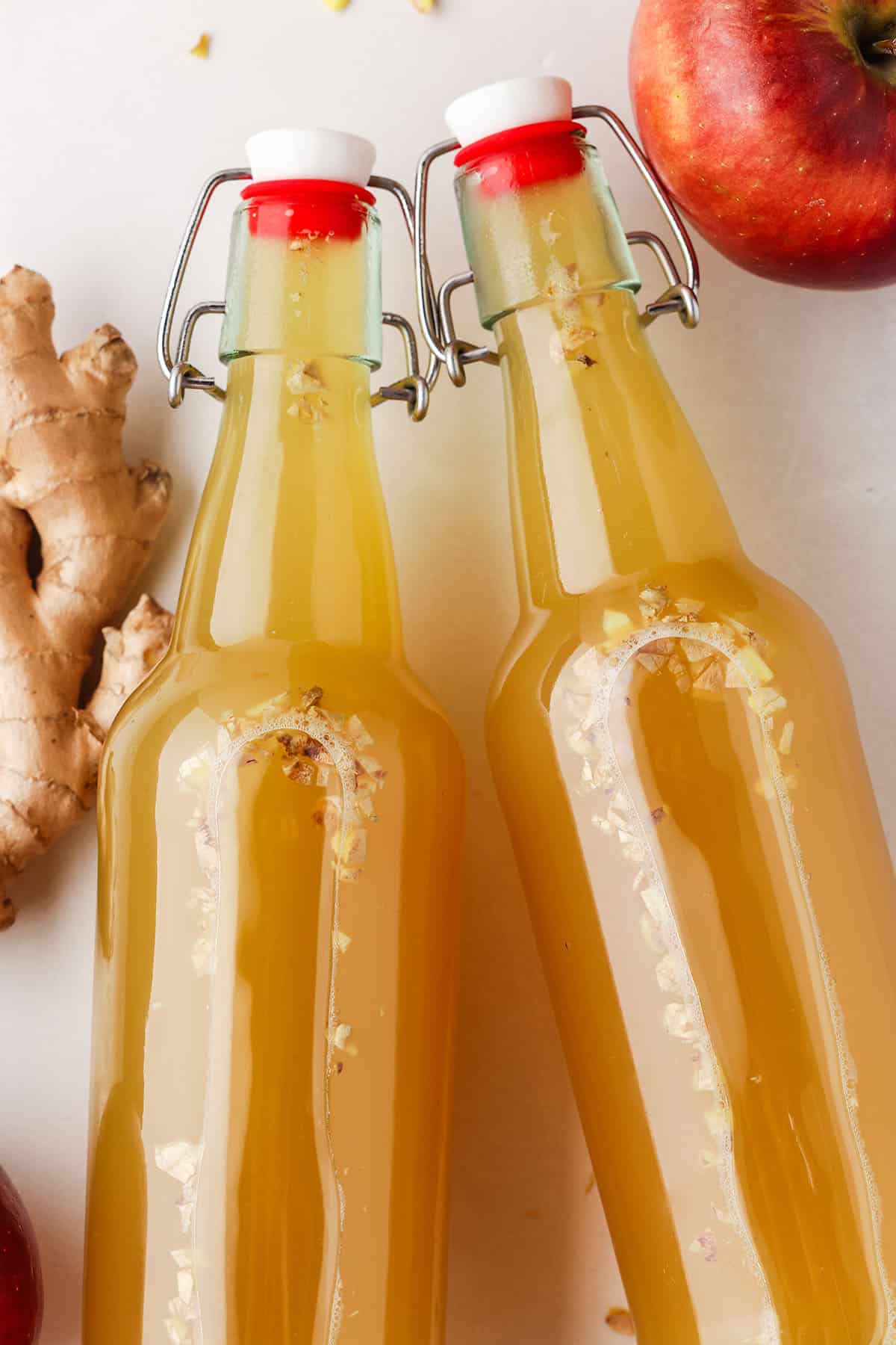 2 bottles of apple ginger kombucha on their sides with a fresh apple and fresh ginger root surrounding. 